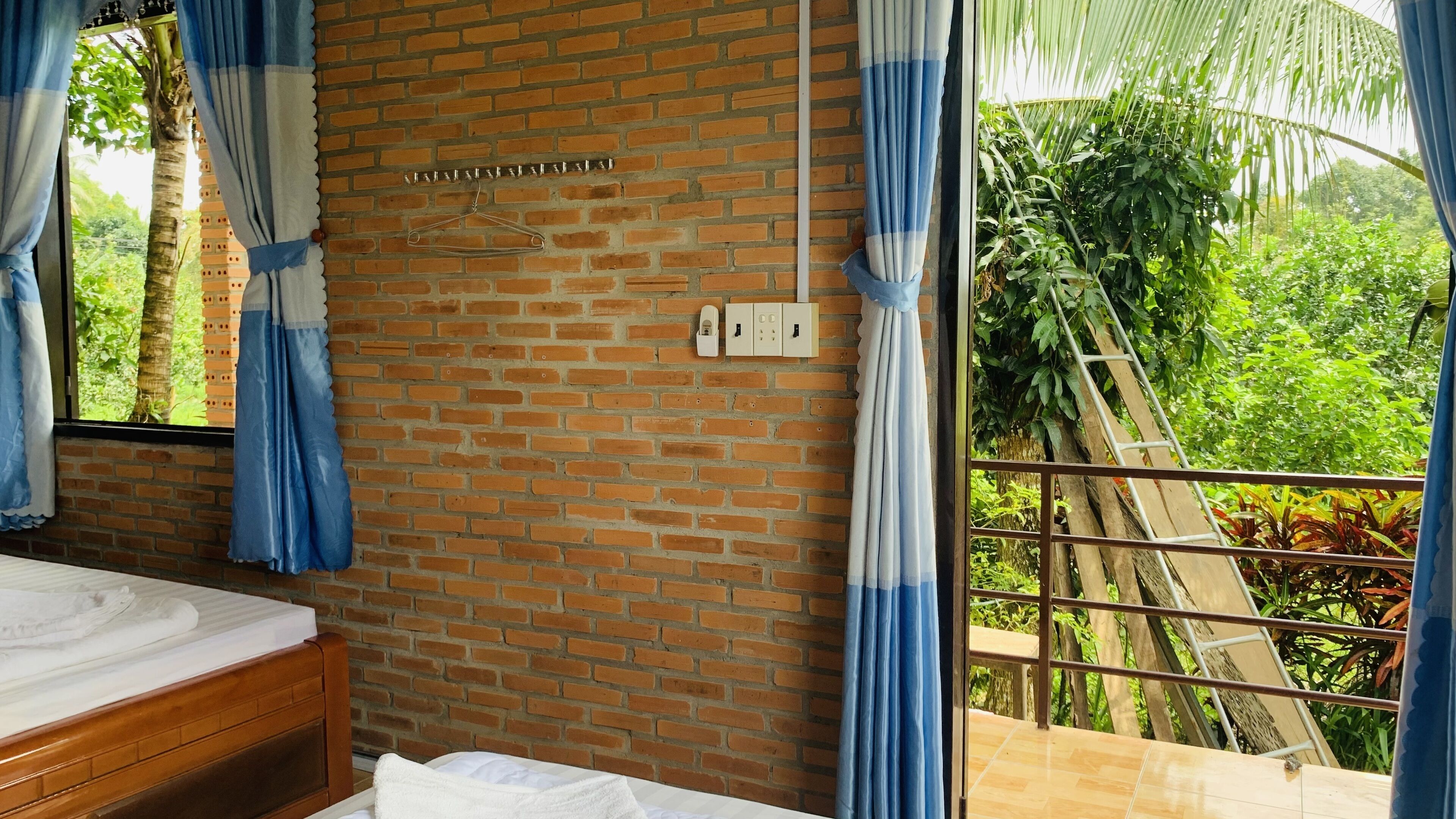 A hotel room with a brick wall, a bed with white linens, a window with blue curtains, and a view of greenery outside.