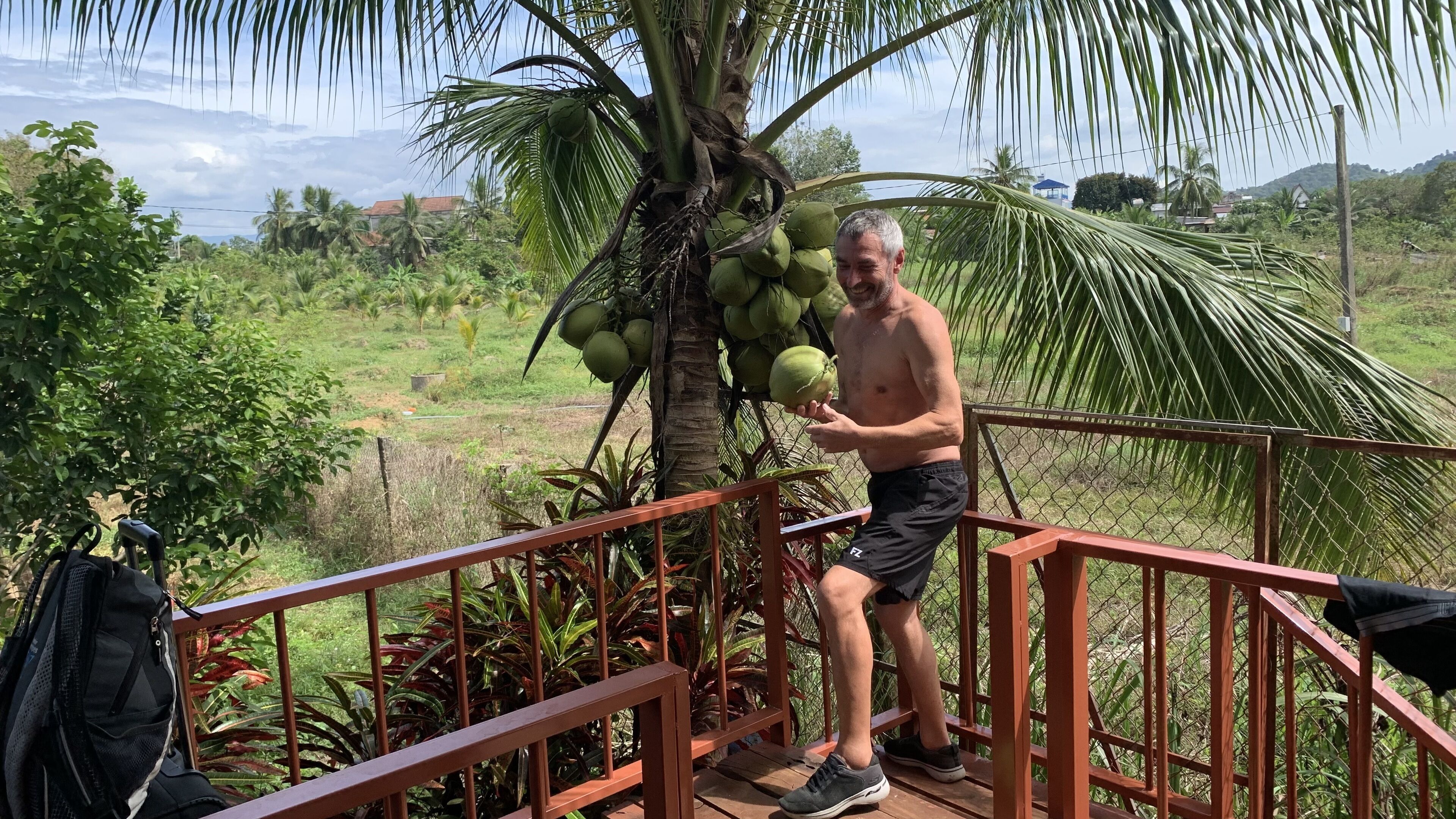 A man standing on a wooden deck with a palm tree and coconuts in the background.