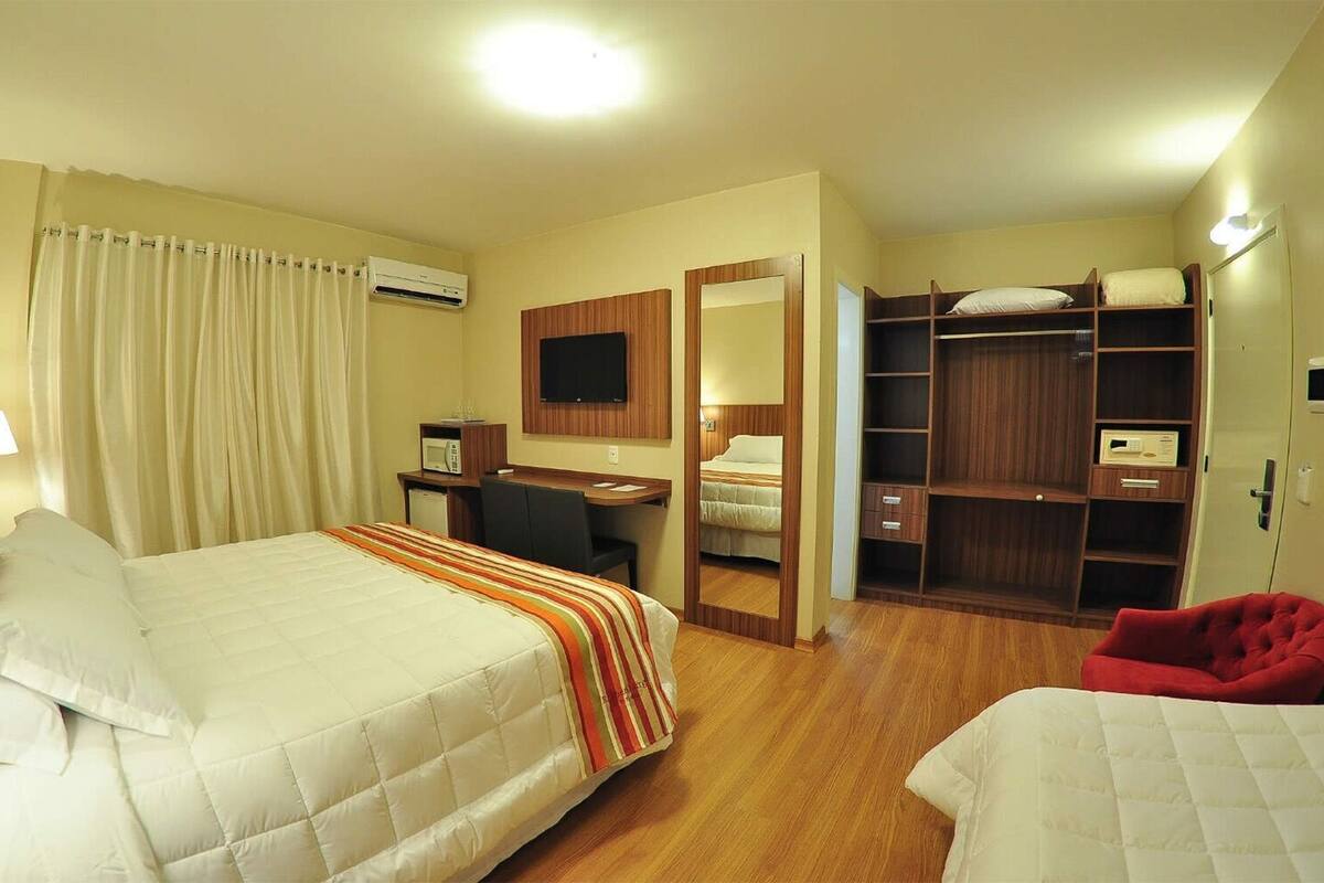 A hotel room with a bed, desk, chair, wardrobe, and a red chair.