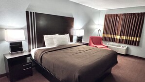 A hotel room with a large bed, two bedside tables with lamps, a red chair, a wall-mounted heater, and a window with curtains. - Owensville Motor Inn (Owensville)