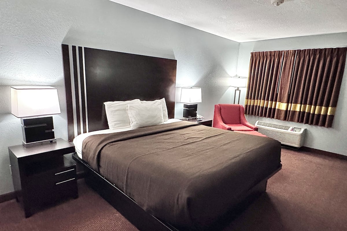 Une chambre d’hôtel avec un grand lit, deux tables de chevet avec des lampes, un fauteuil rouge, un radiateur fixé au mur et une fenêtre avec des rideaux.