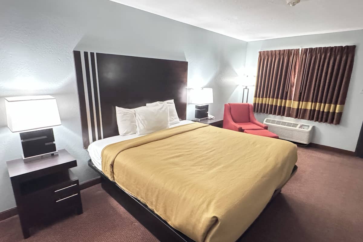 Une chambre d’hôtel avec un lit, des tables de chevet, un fauteuil rouge, une lampe et un appareil de climatisation.