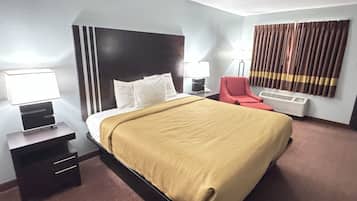 A hotel room with a bed, bedside tables, a red chair, a lamp, and an air conditioning unit.