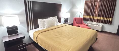 A hotel room with a bed, bedside tables, a red chair, a lamp, and an air conditioning unit.