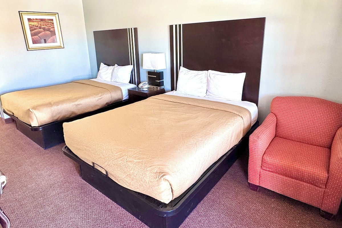 Une chambre d’hôtel avec deux lits, une table de chevet, une lampe, une chaise rouge et un tableau encadré au mur.