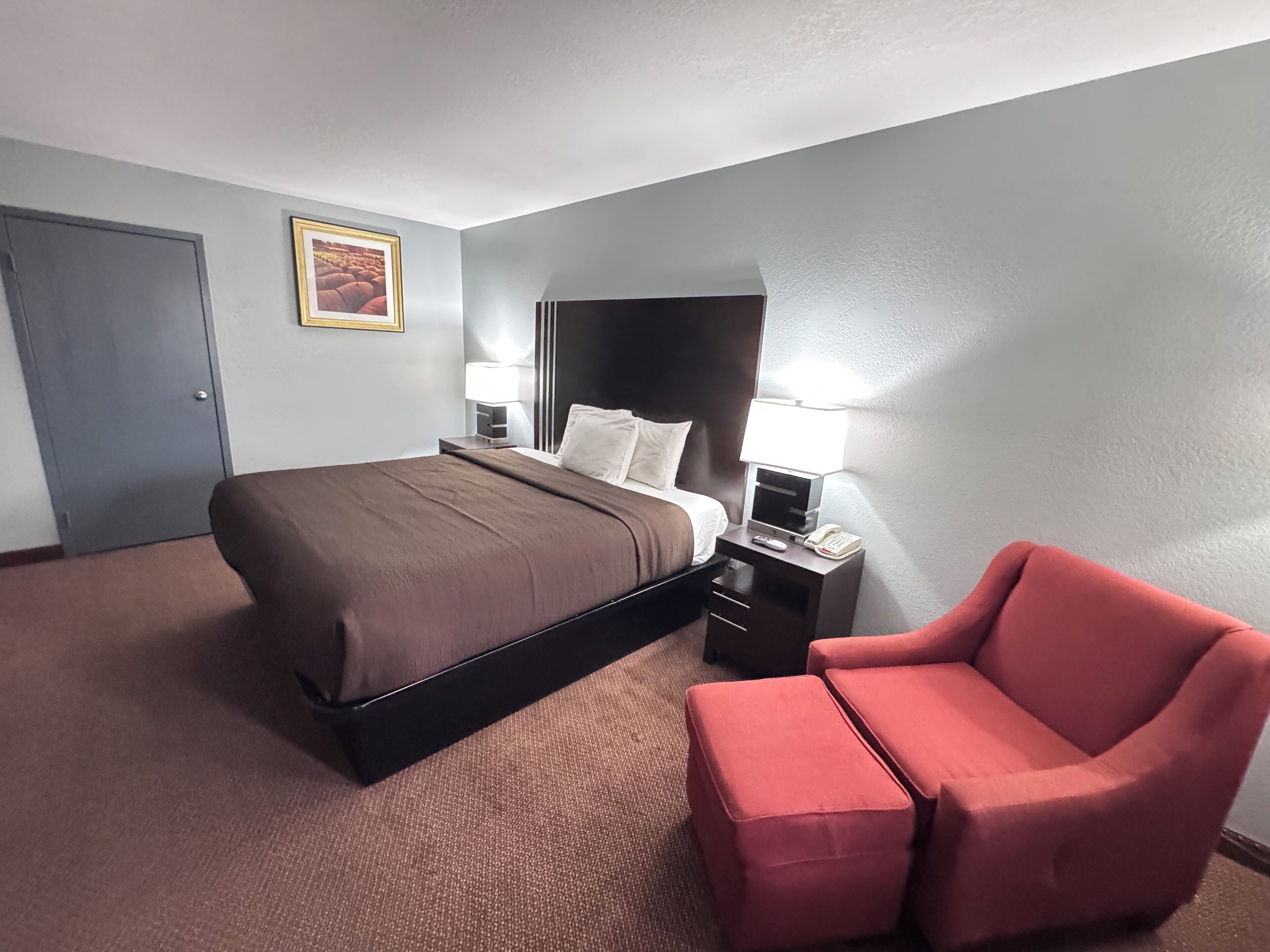 Habitación de hotel con cama, mesitas de noche, una silla, una puerta y un cuadro en la pared.