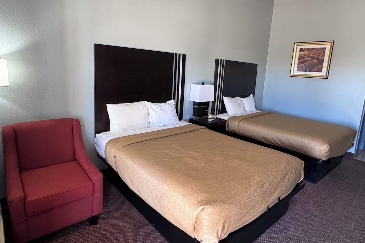 Une chambre d’hôtel avec deux lits, un fauteuil rouge, une table de chevet avec une lampe et un tableau encadré au mur.