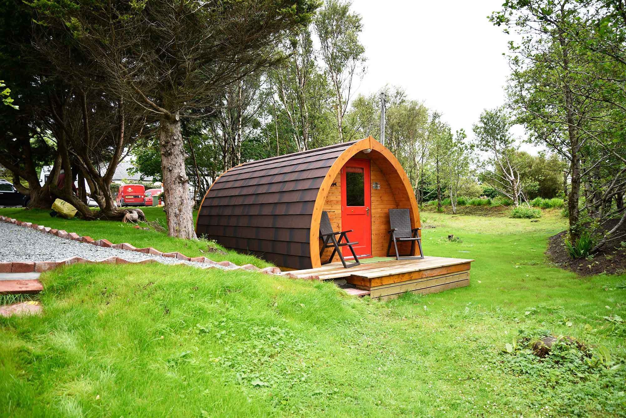A small, wooden, semi-circular cabin with a red door, two black chairs, and a small table on a grassy area surrounded by trees.