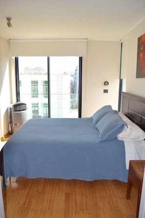 Quarto com uma cama grande, janela com vista para prédios, ventilador de teto e piso de madeira.