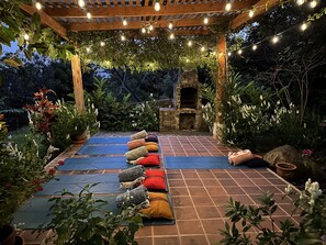 A covered outdoor patio with a stone-tiled floor, a wooden pergola with string lights, and a stone-built pizza oven.