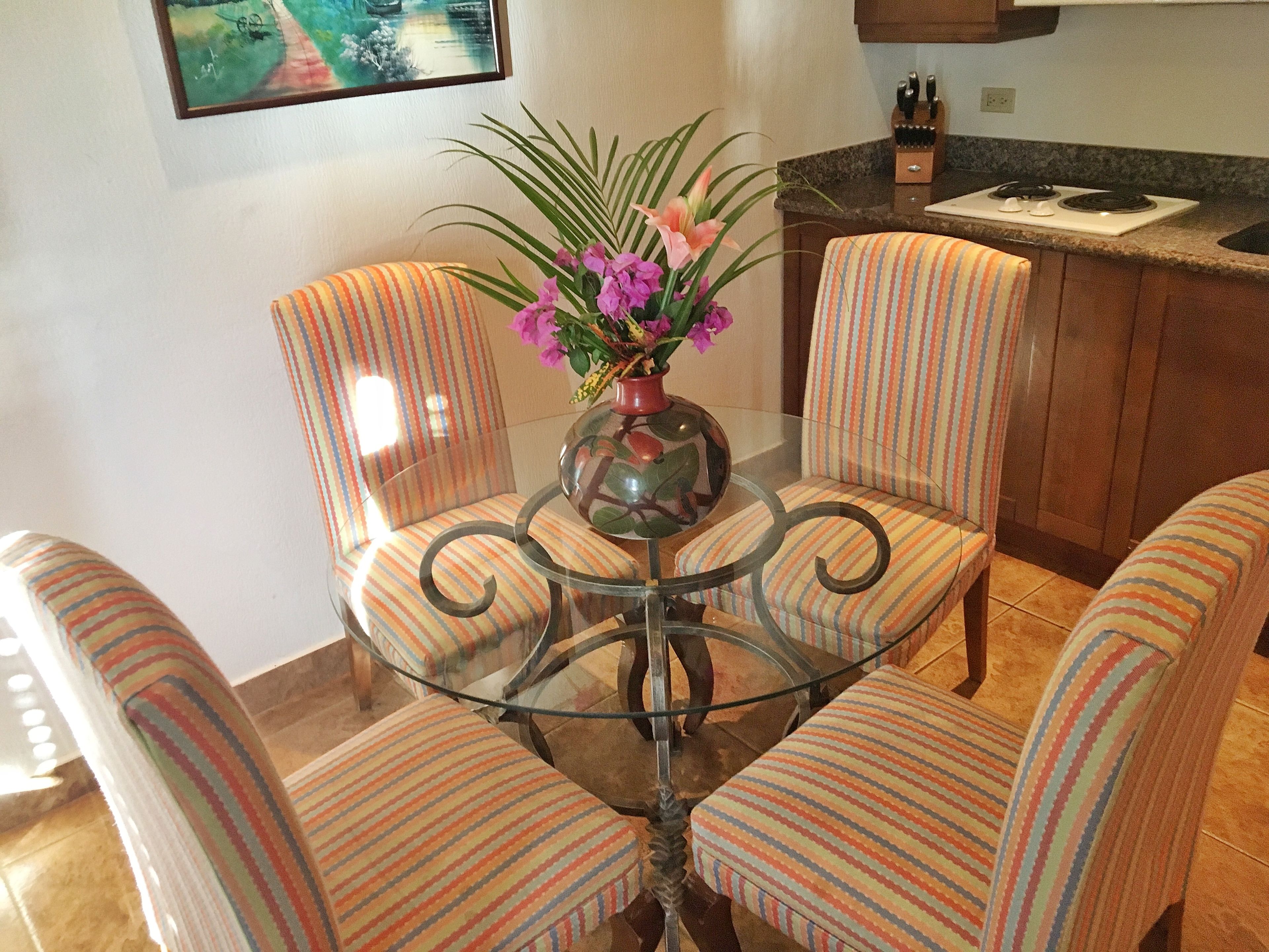A dining area with a glass table surrounded by striped chairs, a vase with flowers, and a kitchen in the background.
