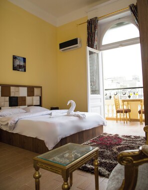 Quarto de hotel com cama grande, uma mesinha, varanda com vista e janela com cortinas.