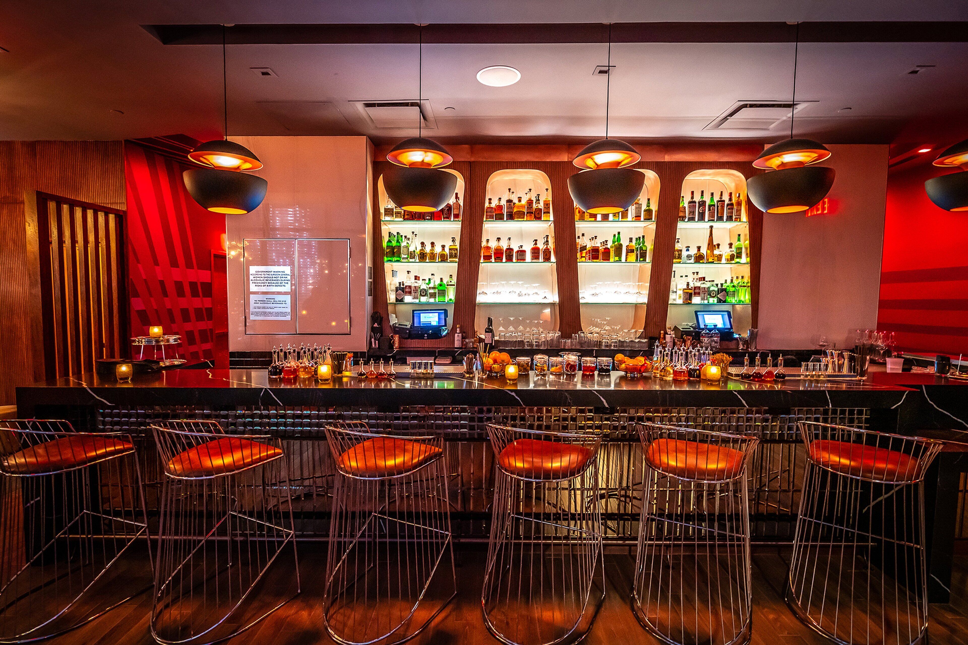 A bar area with multiple stools, shelves of bottles, and hanging pendant lights.