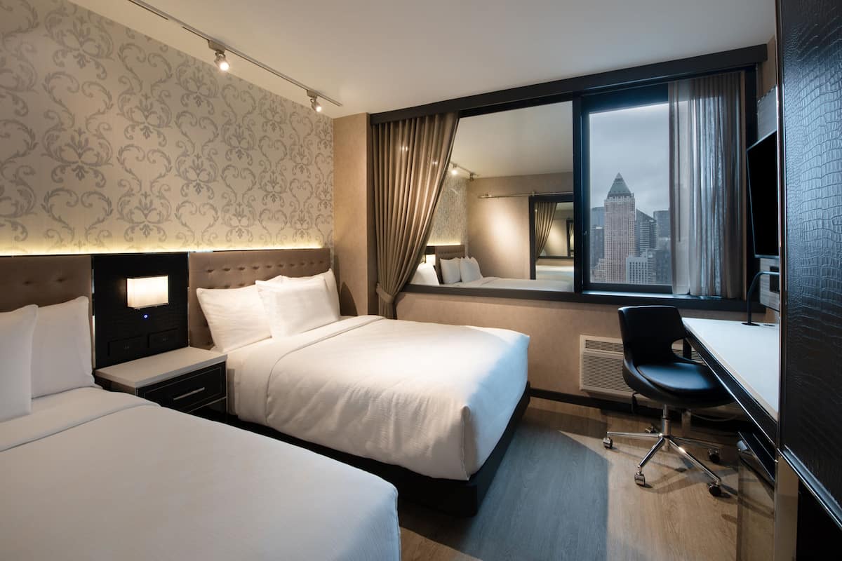 Camera d'albergo con due letti, una scrivania, una sedia, un'ampia finestra con vista sulla città e uno specchio.