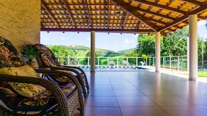 Un espacio de descanso techado al aire libre con mobiliario de mimbre y vistas a una piscina y follaje.