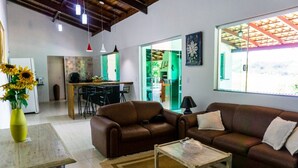 Una sala de estar con un sofá de cuero marrón, una mesa de centro de madera y un jarrón con girasoles. Se observa una cocina al fondo con una mesa de comedor y sillas.
