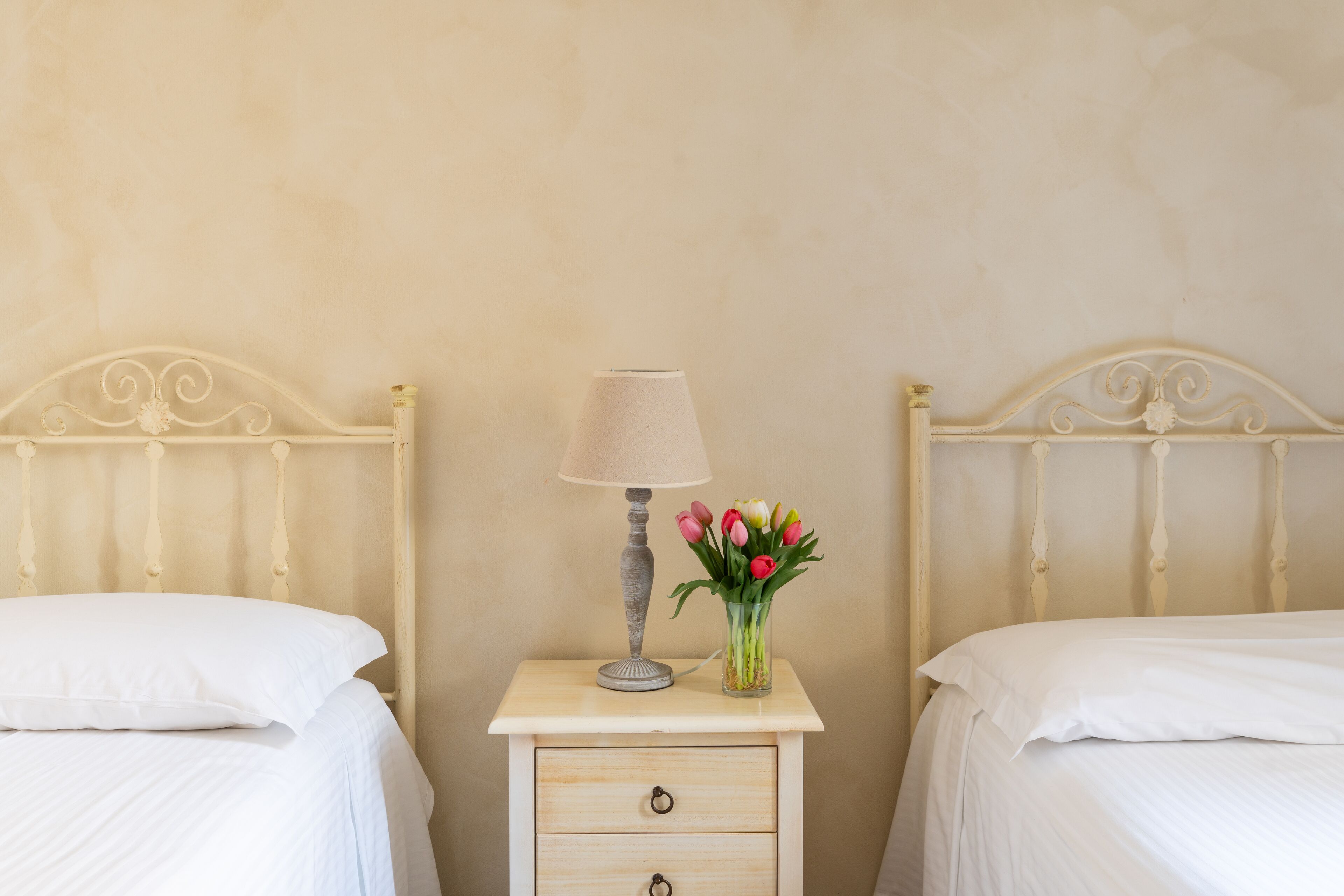 Deux lits simples avec du linge de lit blanc, une table de chevet avec une lampe et un vase de fleurs, et un mur de couleur claire.