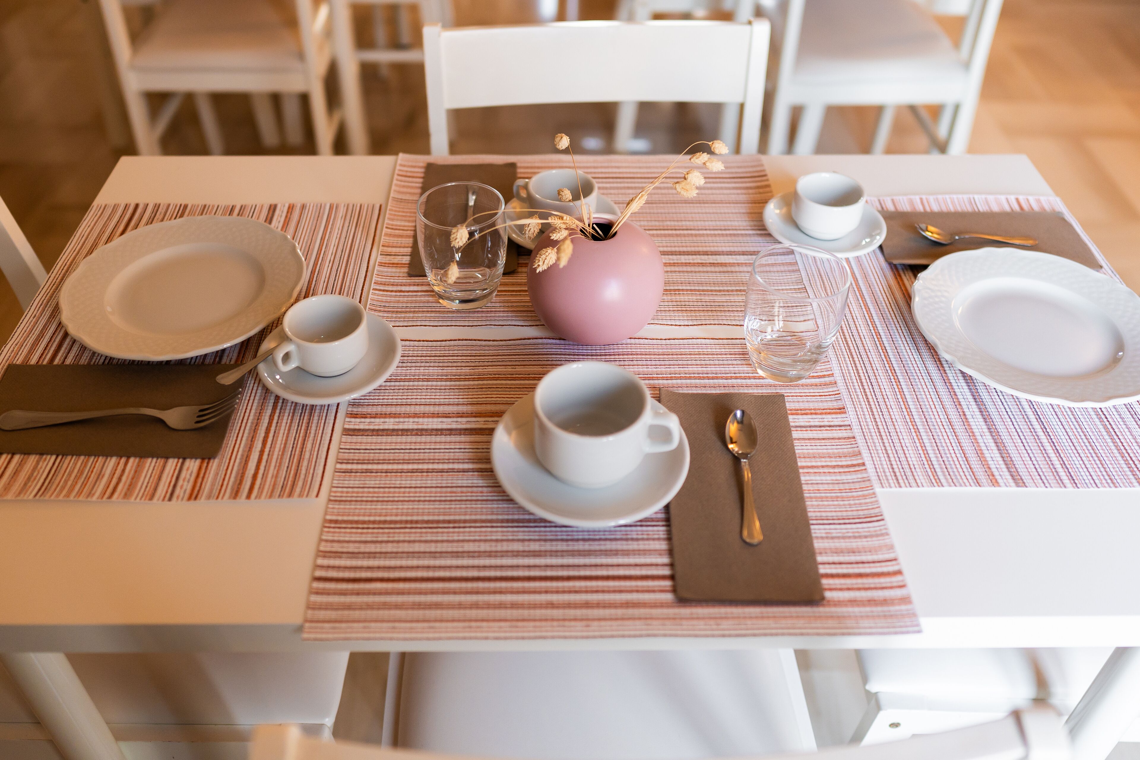 A neatly set dining table with plates, cups, and cutlery, a vase with dried flowers, and a striped tablecloth.