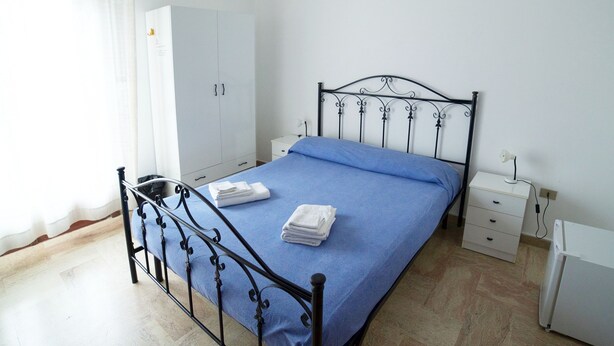 A bedroom with a metal bed frame, a blue bedspread, white towels, a white wardrobe, a bedside table with a lamp, and a radiator. - Gallipolis Terra (Gallipoli)