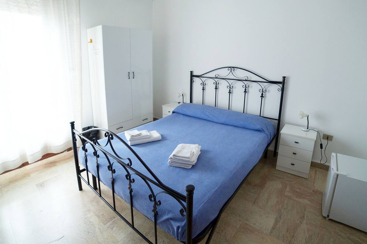 Un dormitorio con estructura de cama metálica, cubrecama azul, toallas blancas, armario blanco, mesita de noche con lámpara y un radiador.