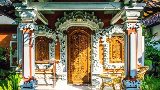 Un bâtiment traditionnel doté de portes et de colonnes en bois richement décorées, d’un sol carrelé et d’un petit espace extérieur aménagé avec des chaises en osier.