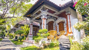 Un bâtiment traditionnel, doté d’un toit en tuiles, de colonnes décorées et d’un jardin bien entretenu, agrémenté de plantes en pot et d’un sentier.