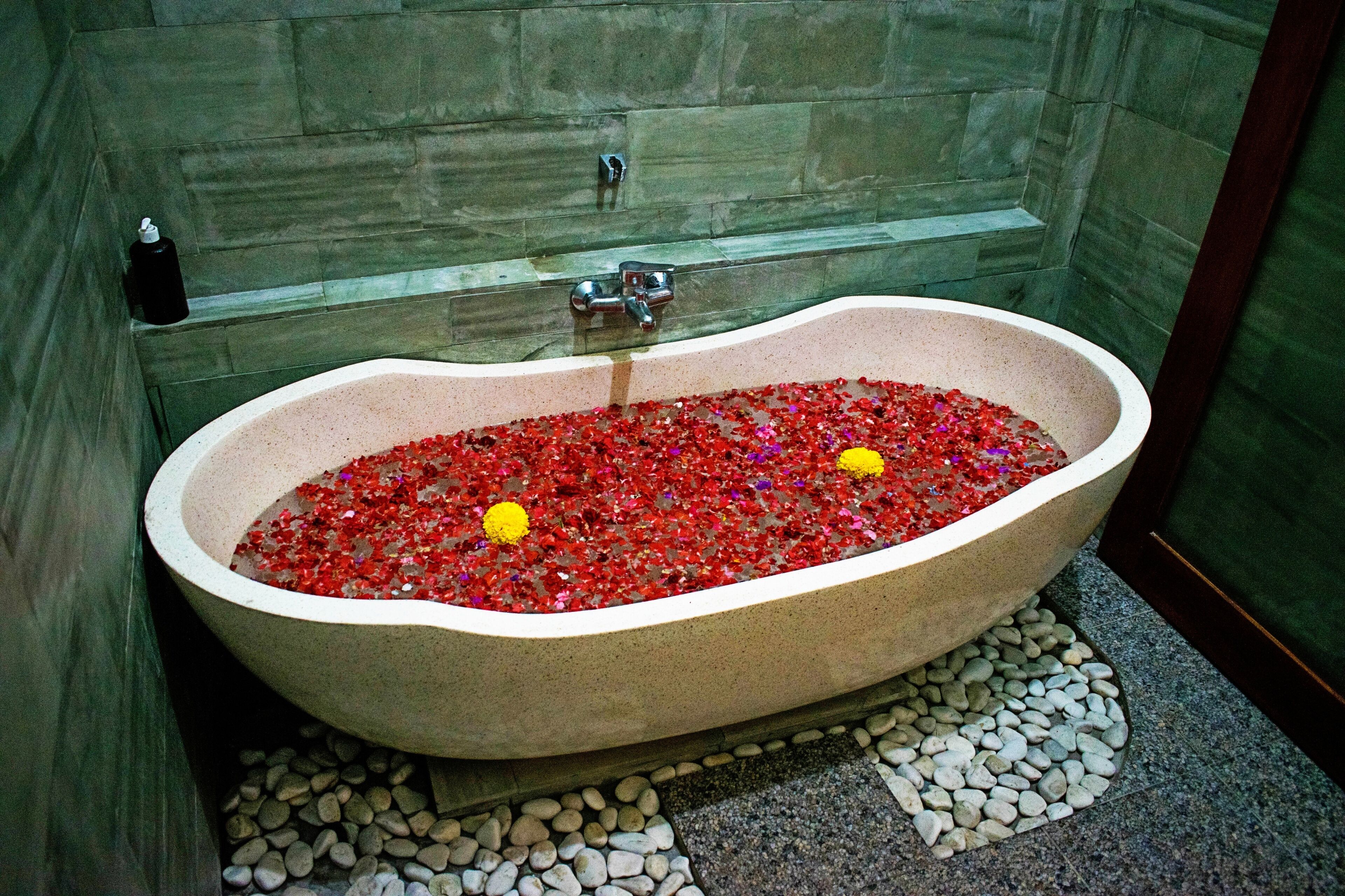 Una bañera llena de guijarros rojos y flores amarillas, ubicada en un baño con paredes y piso de piedra.
