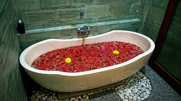 Una bañera llena de guijarros rojos y flores amarillas, ubicada en un baño con paredes y piso de piedra.
