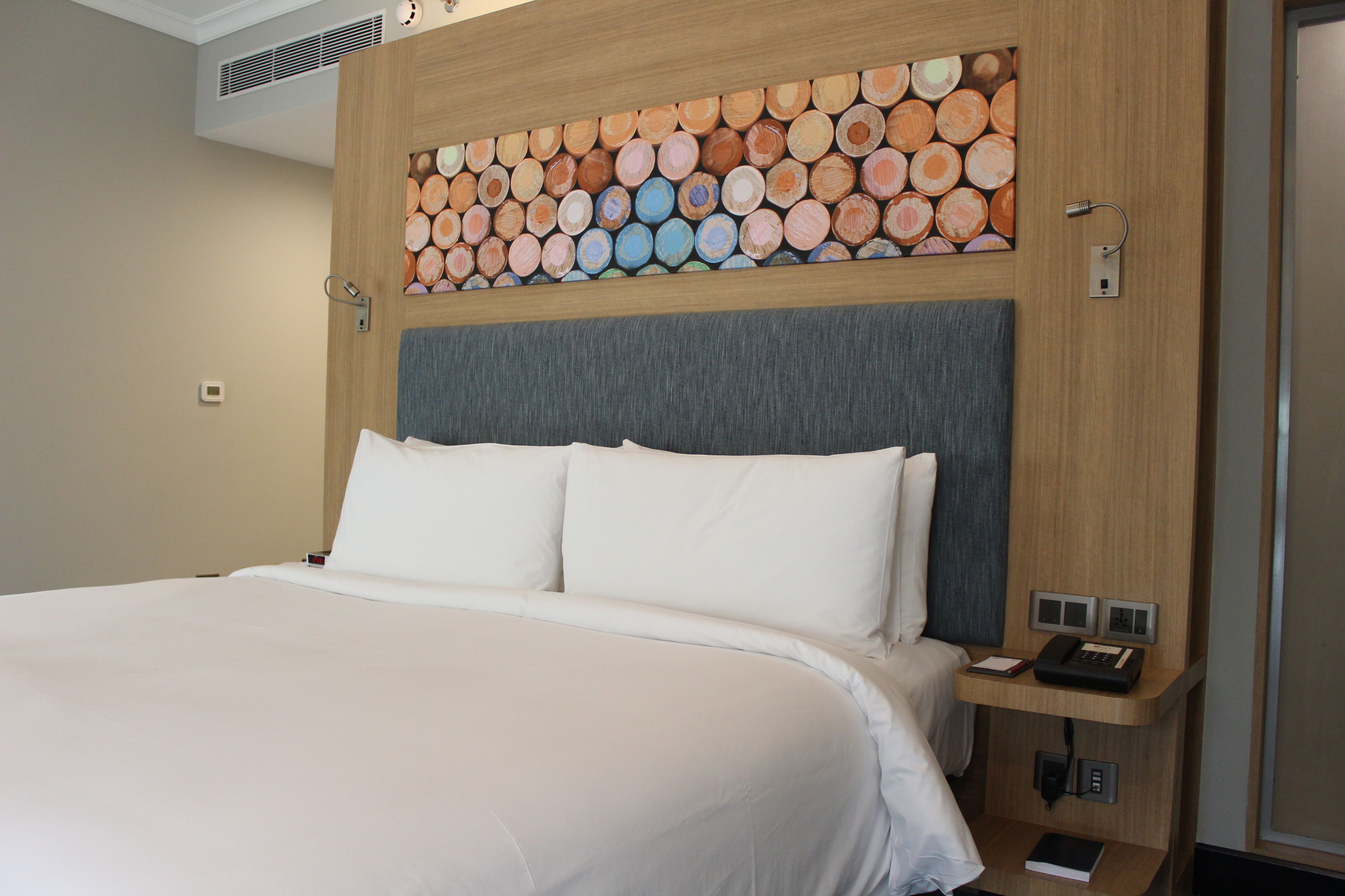 Habitación de hotel con una cama grande, cabecera de madera con un diseño circular, mesita de noche con un teléfono y un libro, y un aire acondicionado de pared.