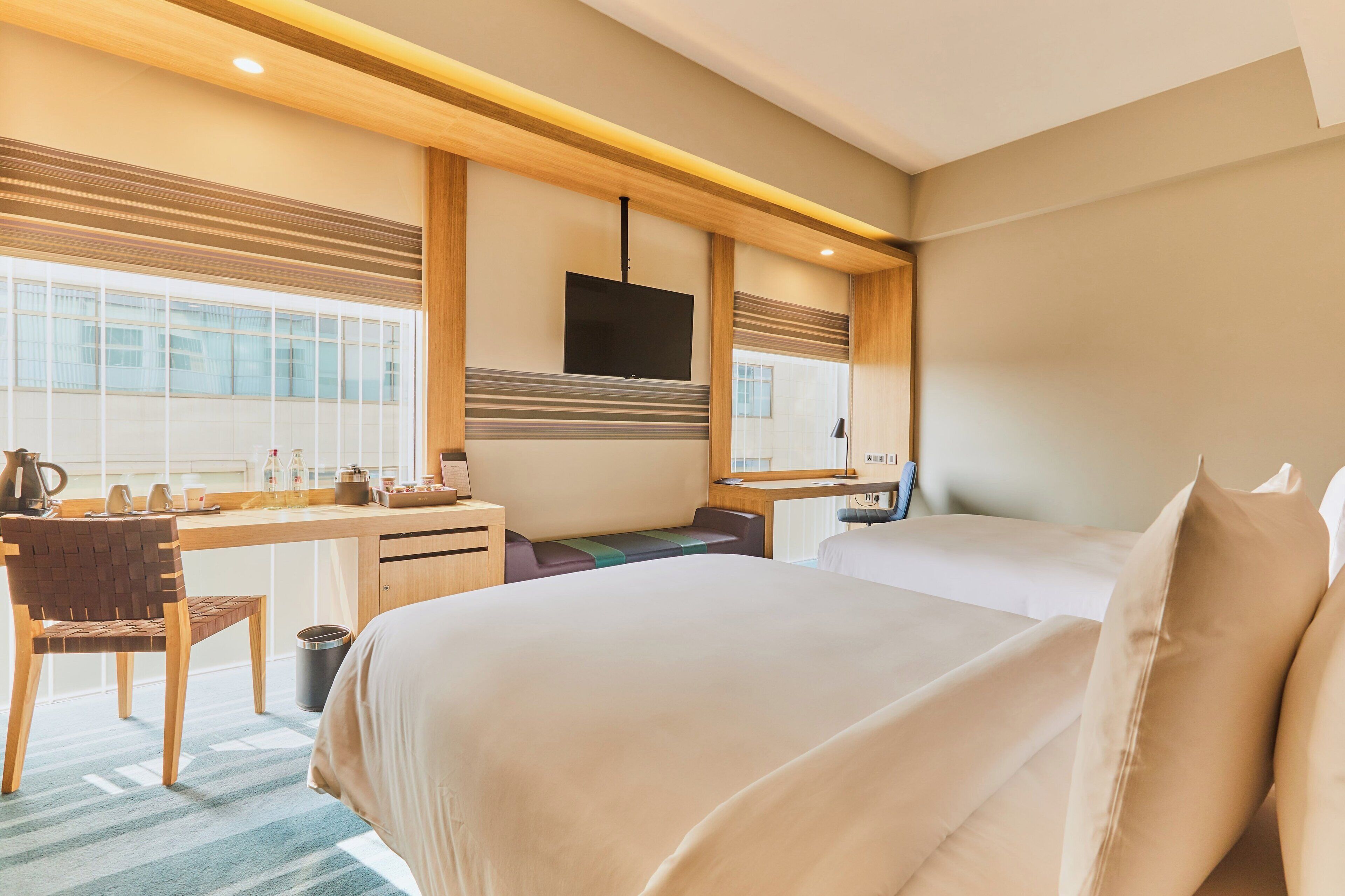Ein Hotelzimmer mit einem großen Bett, einem Schreibtisch mit Stuhl, einem an der Wand montierten Fernseher und einem Fenster mit Jalousien.