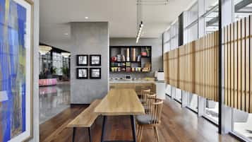 A modern interior with a long wooden table and chairs, a kitchen area with shelves, and large windows.