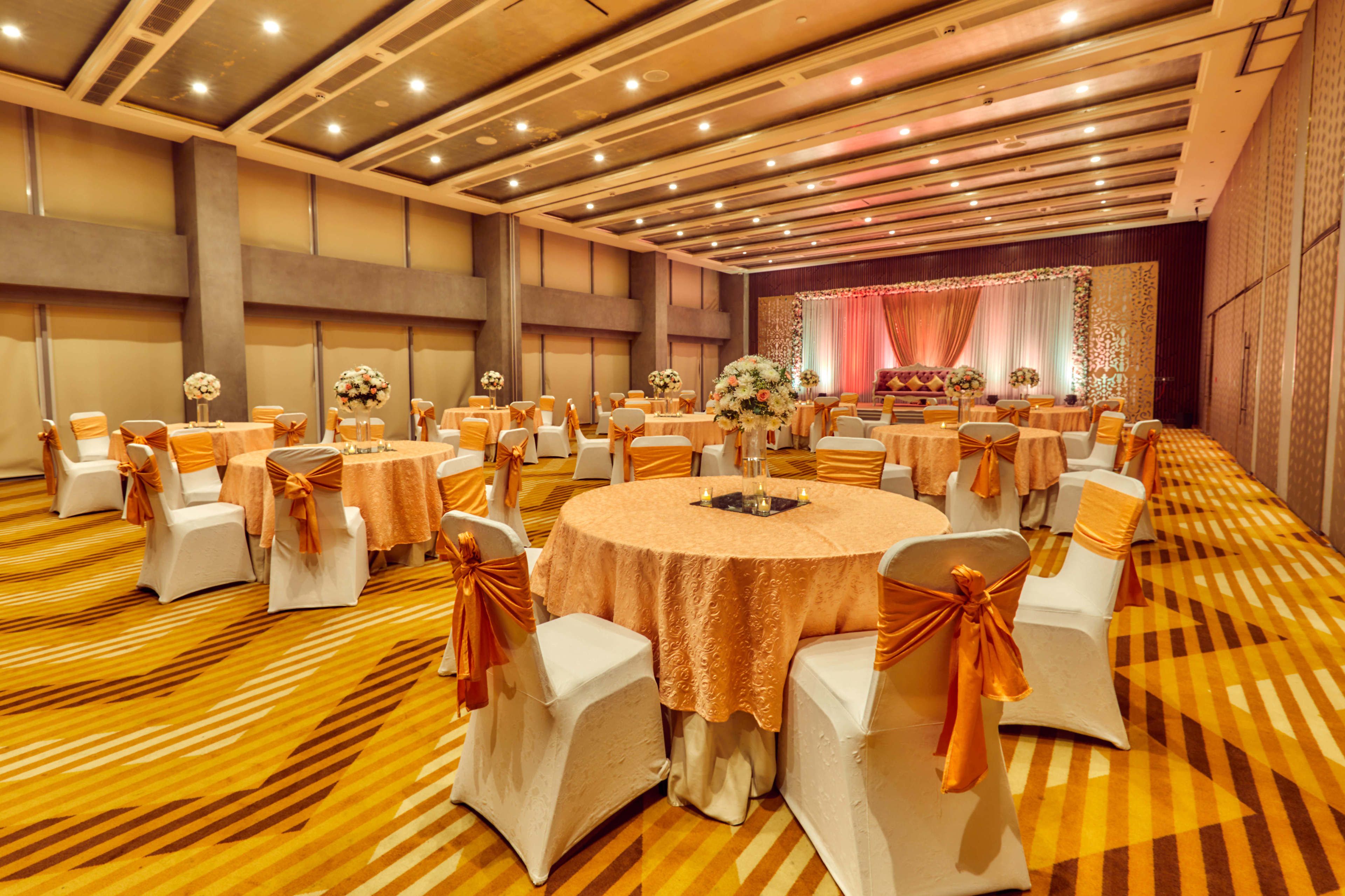 A banquet hall with round tables, white chairs with orange sashes, and a stage with drapes.