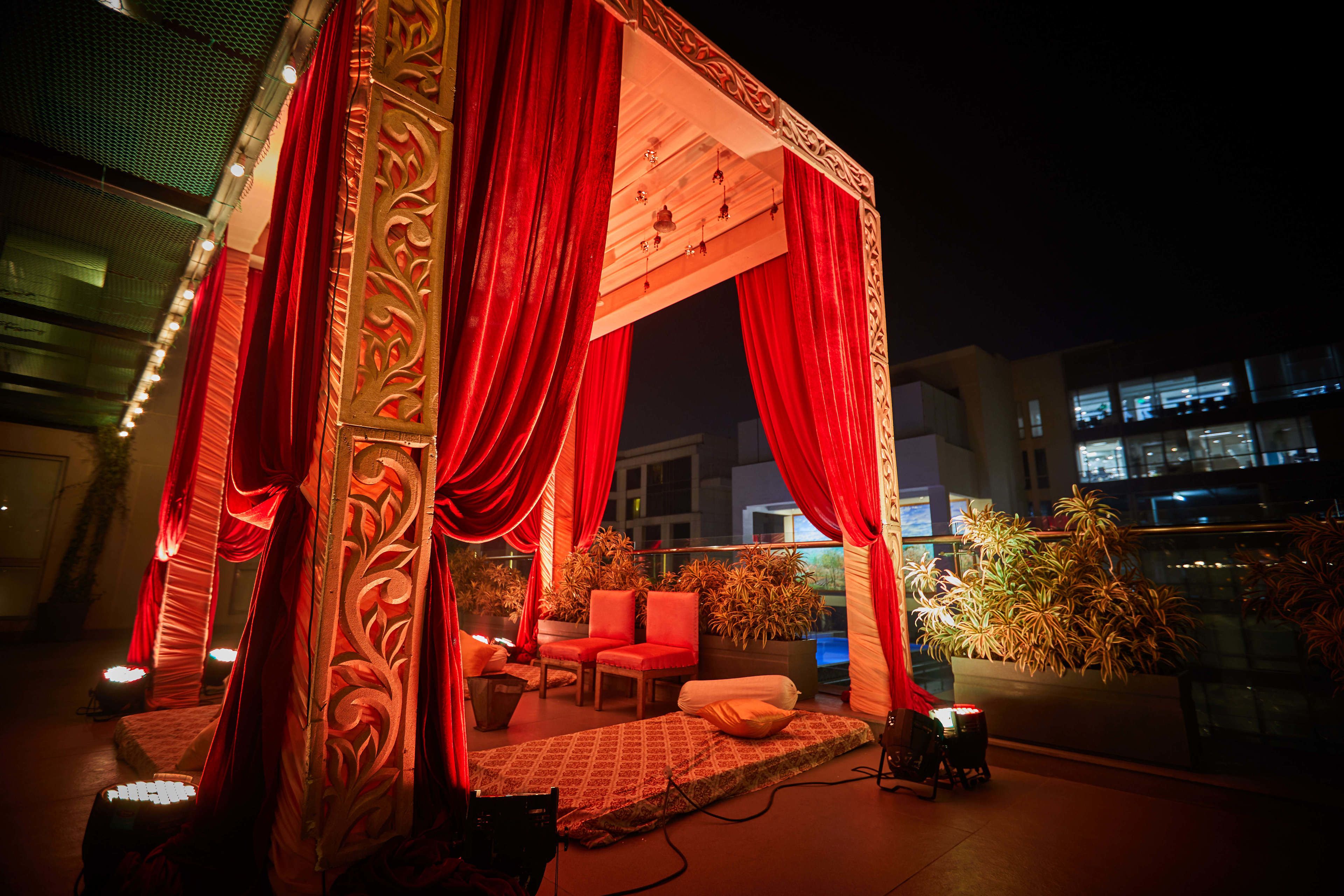 A stage with red curtains and a patterned canopy, surrounded by potted plants and seating.
