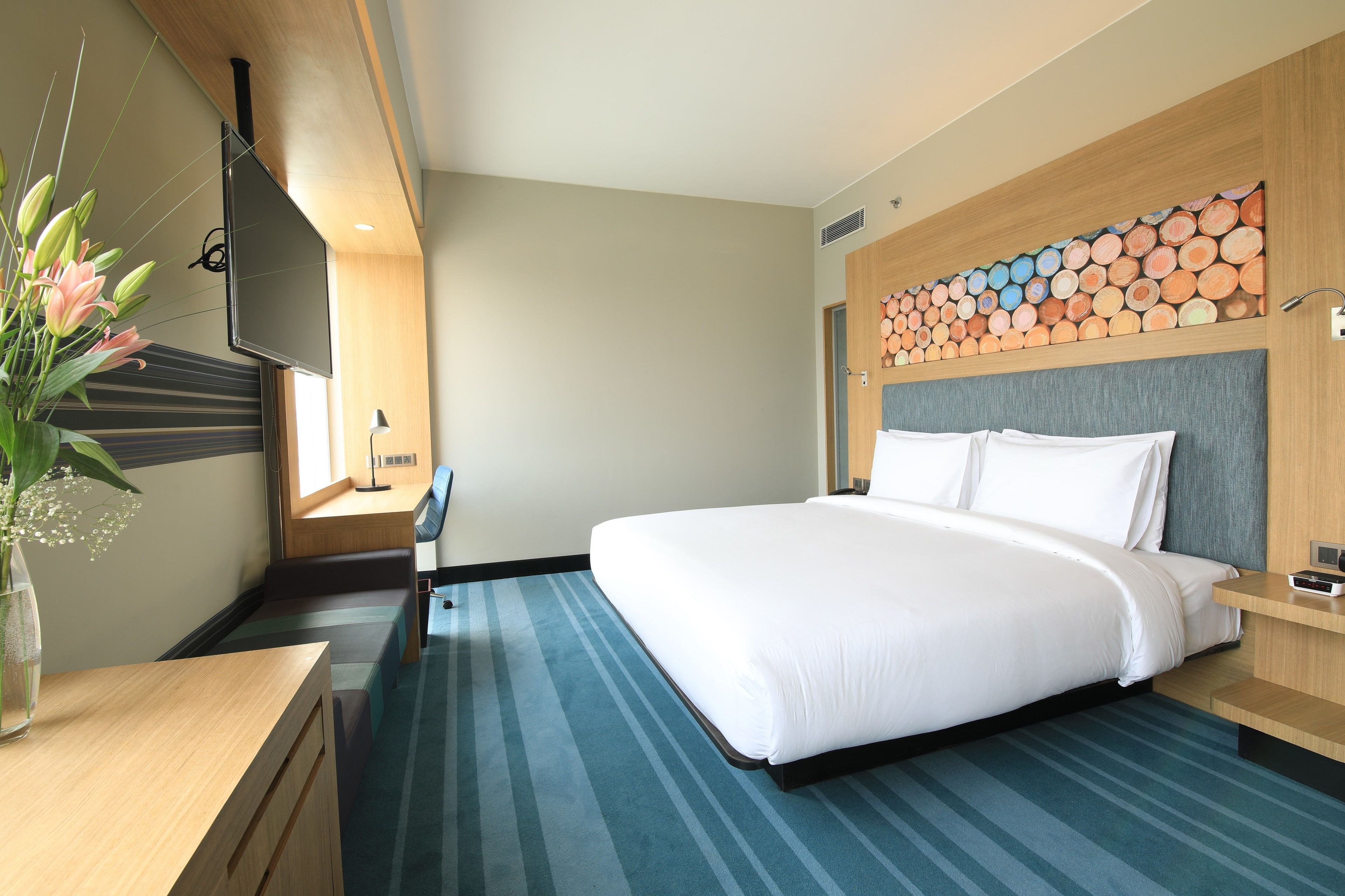 Habitación de hotel con una cama grande, un escritorio, una silla, un televisor montado en la pared y un original cabecero de madera con un diseño de tronco de colores.