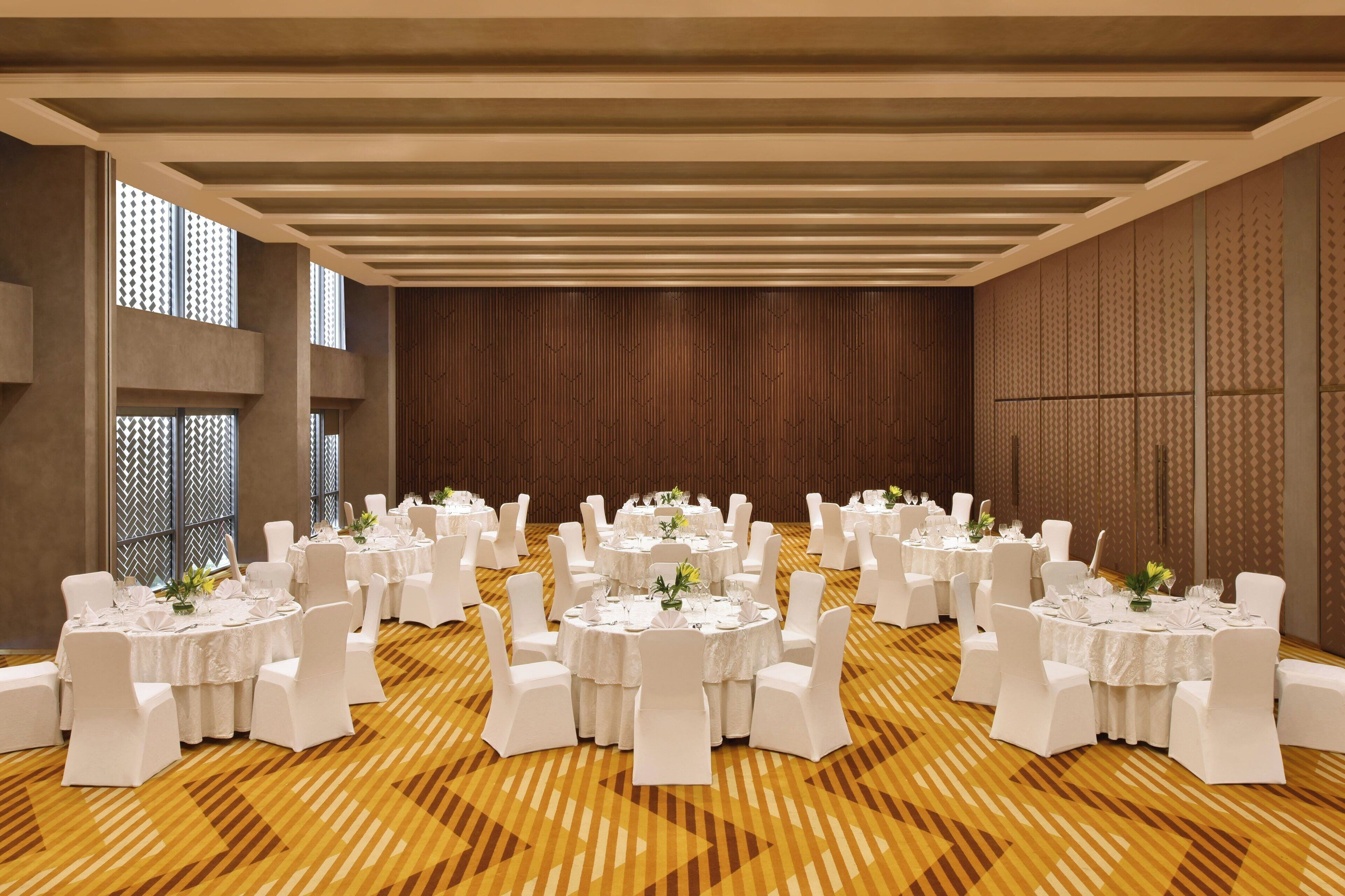 Een feestzaal met meerdere ronde tafels, witte stoelen en een houten vloer met patroon.