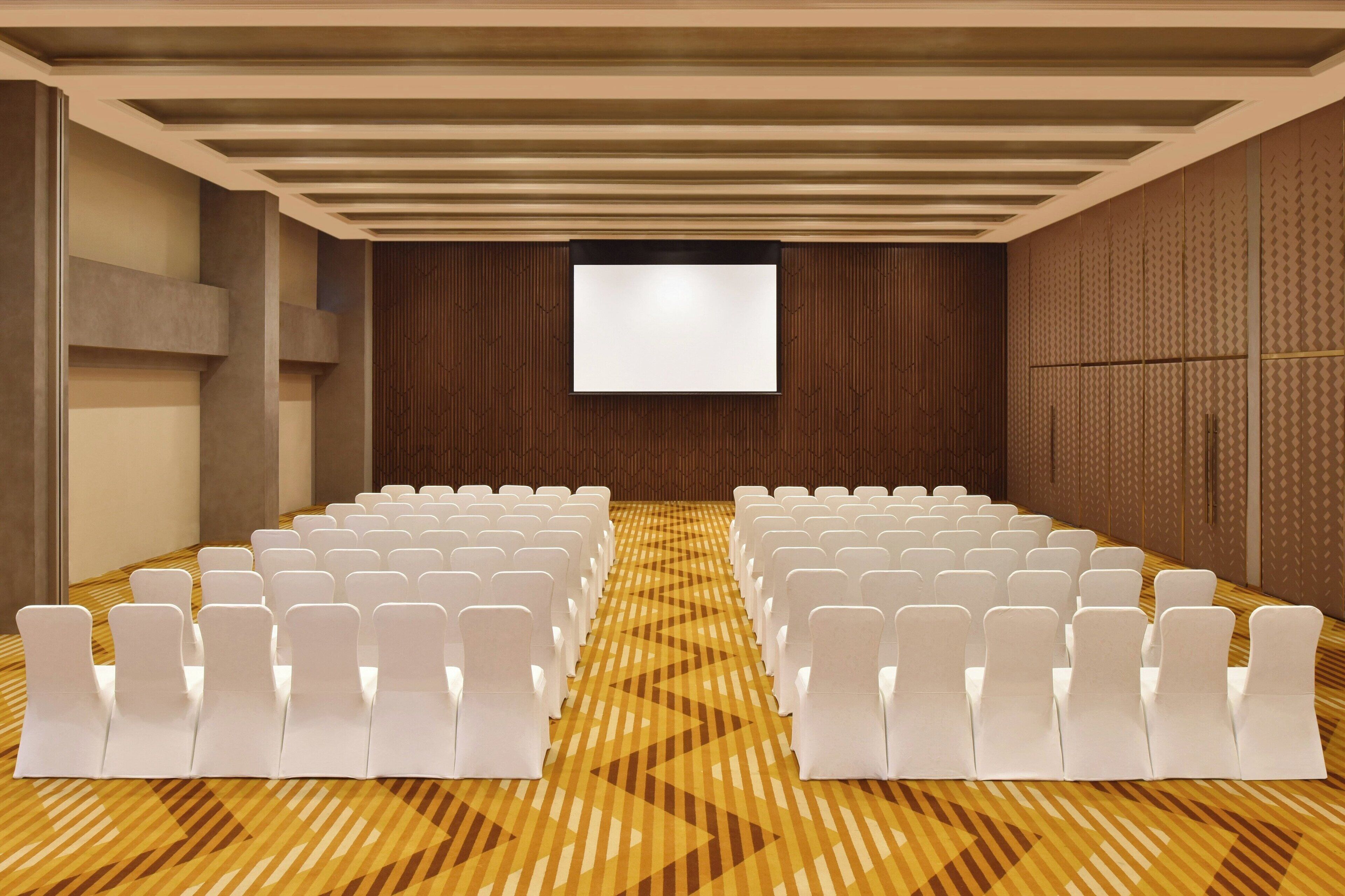 A conference room with rows of white chairs, a large screen, and a patterned carpet.