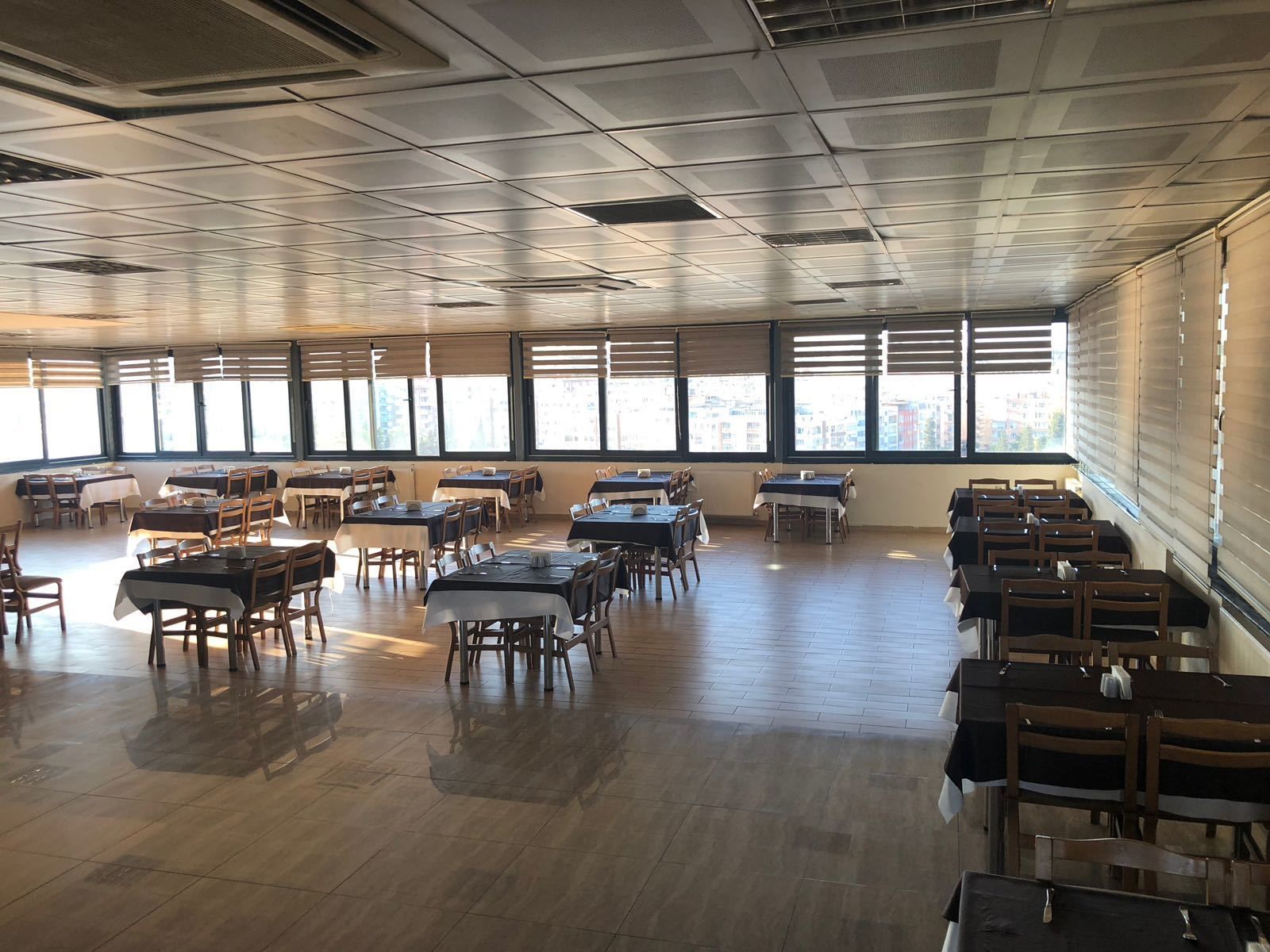 A spacious dining area with multiple tables and chairs, large windows providing natural light, and a high ceiling with ventilation panels.