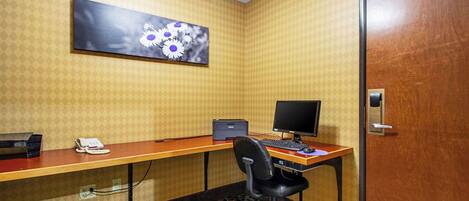 A hotel room with a wooden desk, a computer, a chair, a telephone, and a painting on the wall.