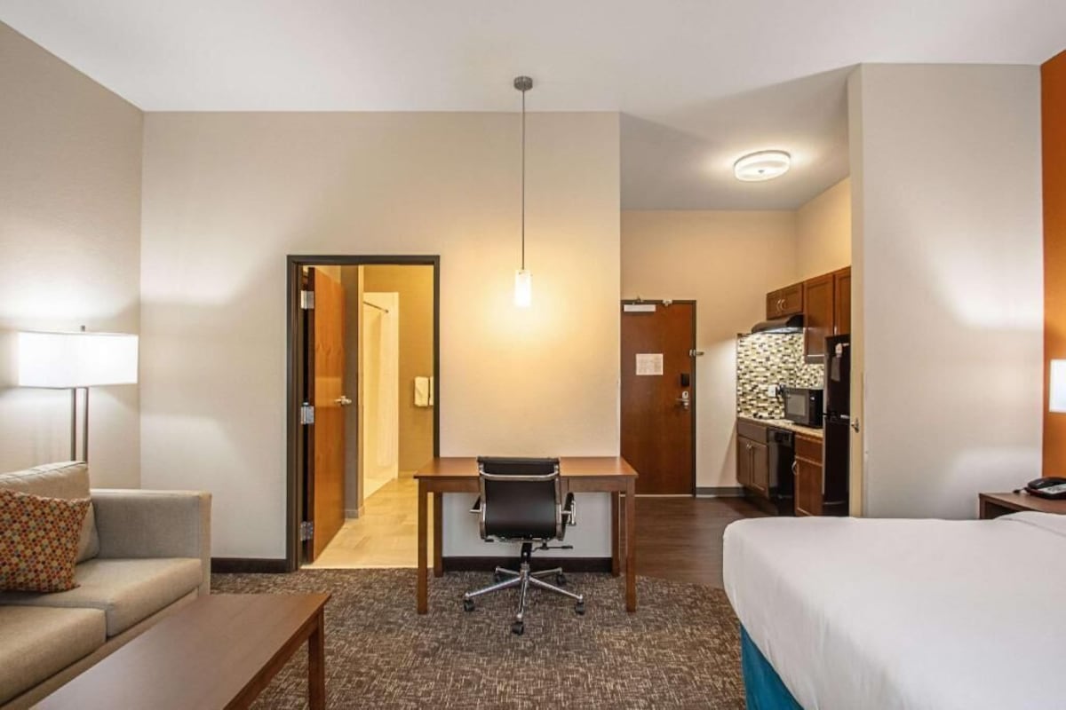 A hotel room with a bed, a desk, a chair, a lamp, a kitchenette, and a door leading to another room.