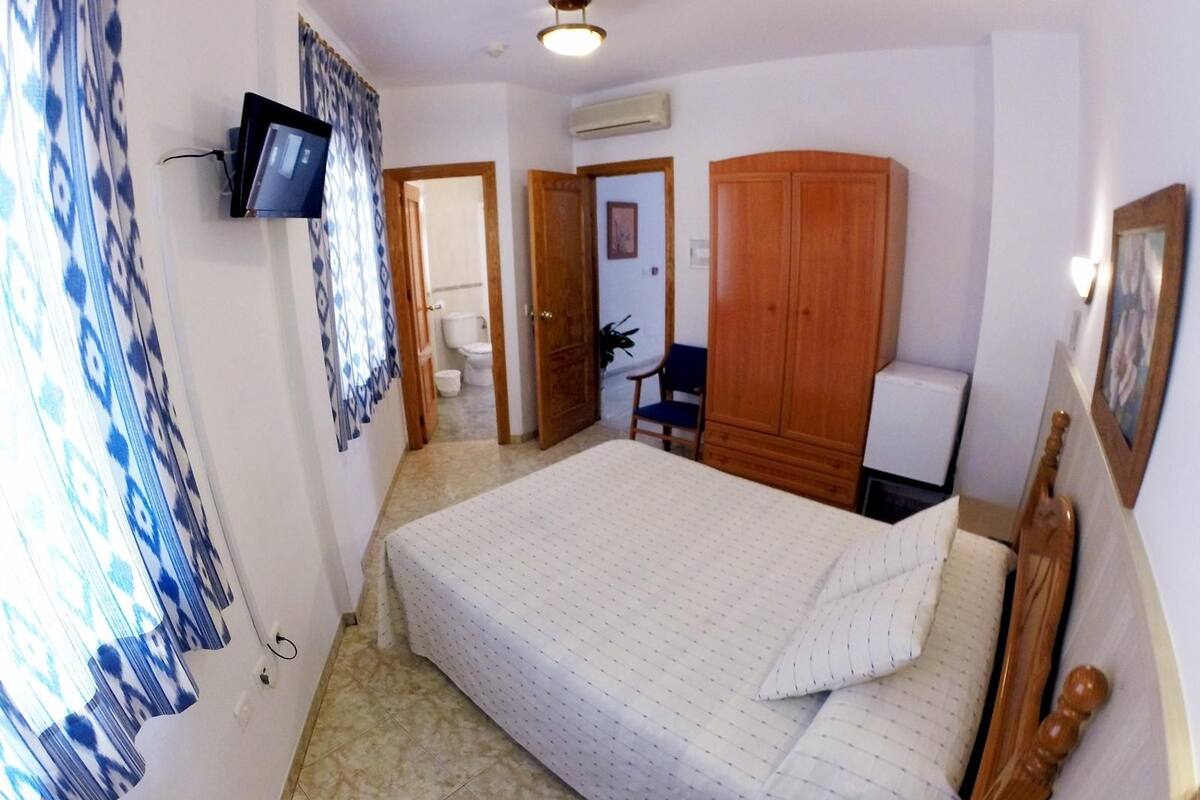 Un dormitorio pequeño y ordenado con una cama, un armario de madera, un televisor montado en la pared y un baño visible a través de una puerta abierta.