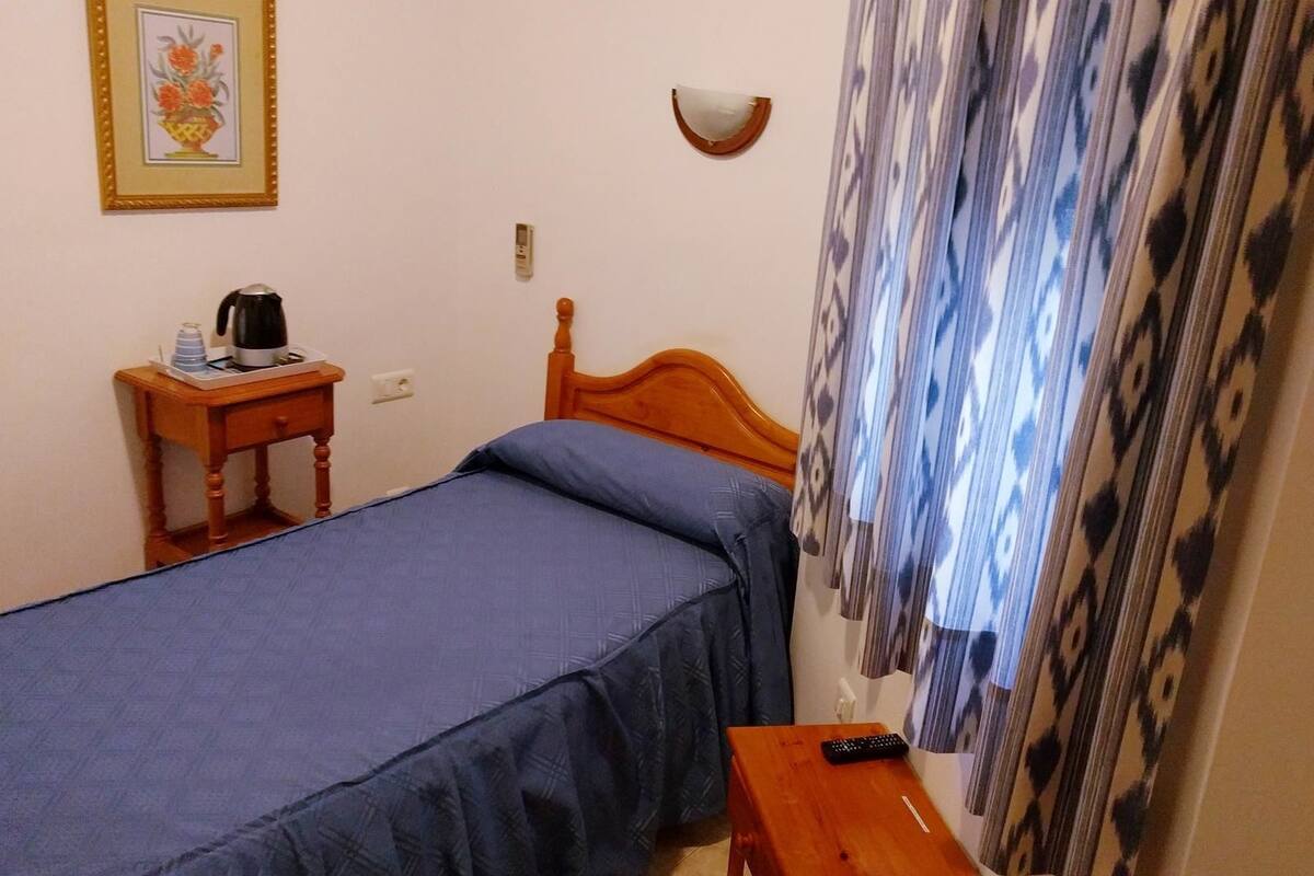 Un dormitorio pequeño y ordenado con una cama individual, una mesita de noche con hervidor, un cuadro enmarcado en la pared y una cortina estampada.