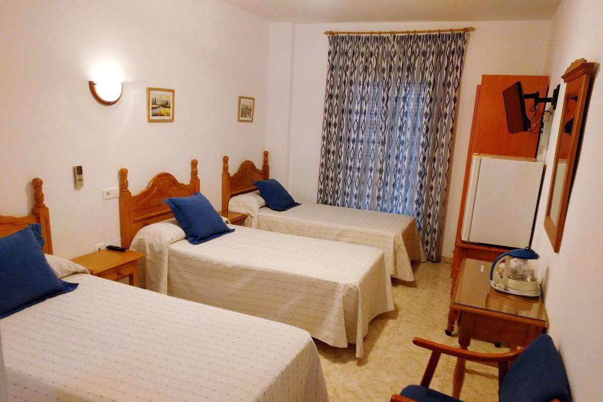 Una habitación de hotel con dos camas individuales, una silla de madera, un escritorio con lámpara, un espejo y un refrigerador.