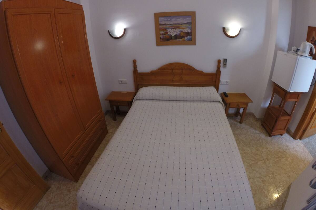 Un dormitorio con una cama de madera, dos mesitas de noche, un armario, un cuadro colgado en la pared y un refrigerador blanco.