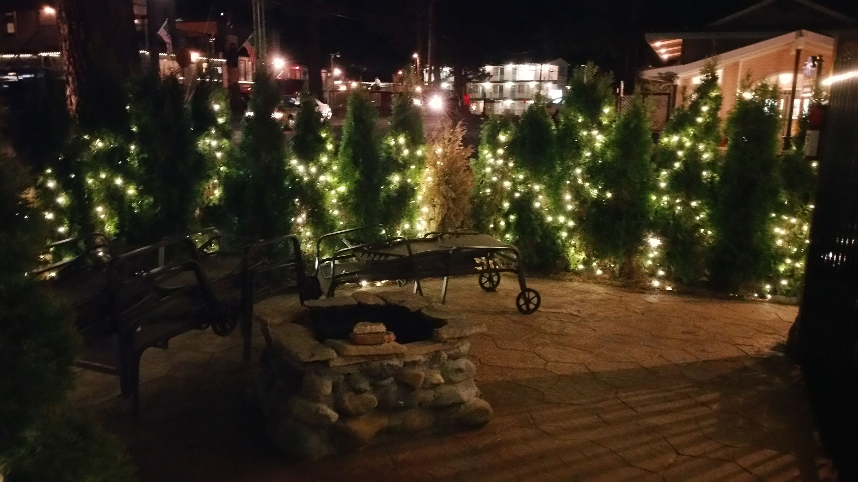 Een buitenruimte 's nachts met kerstbomen versierd met lichtjes, een vuurkorf van steen en een metalen bank.
