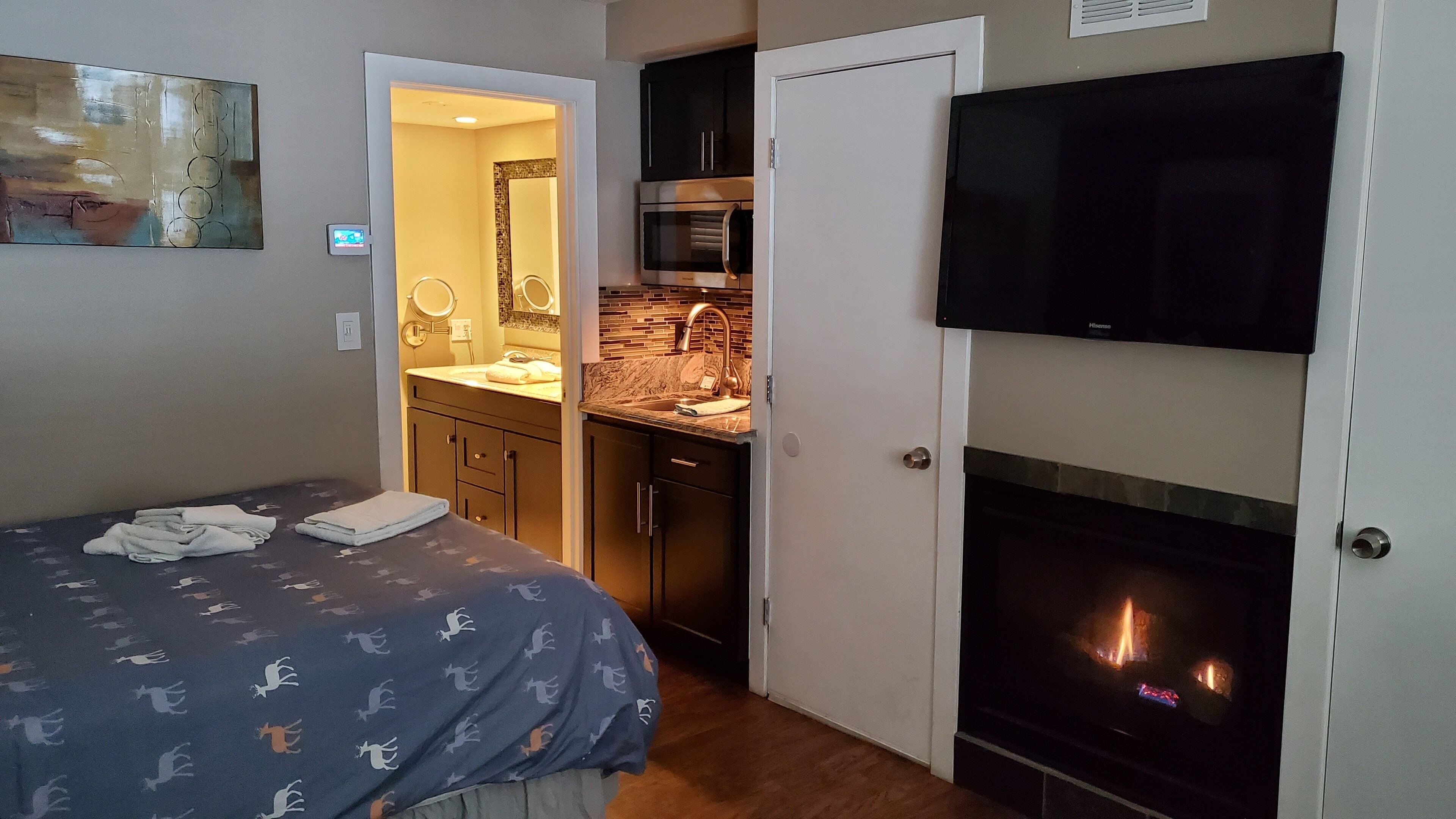 A bedroom with a bed, a TV, a fireplace, and a kitchen area with a microwave and sink.