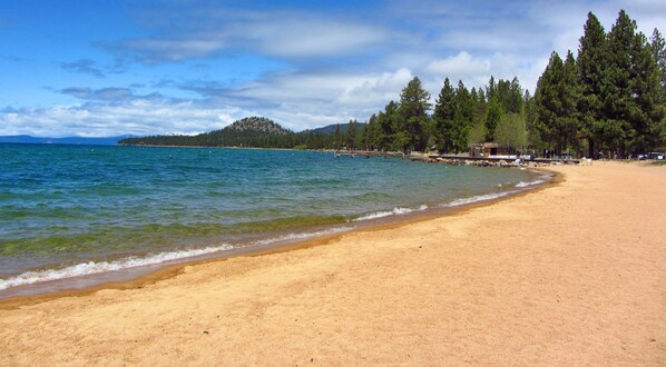 Ein Strand mit klarem Wasser, sandigem Ufer und einem bewaldeten Hügel im Hintergrund.