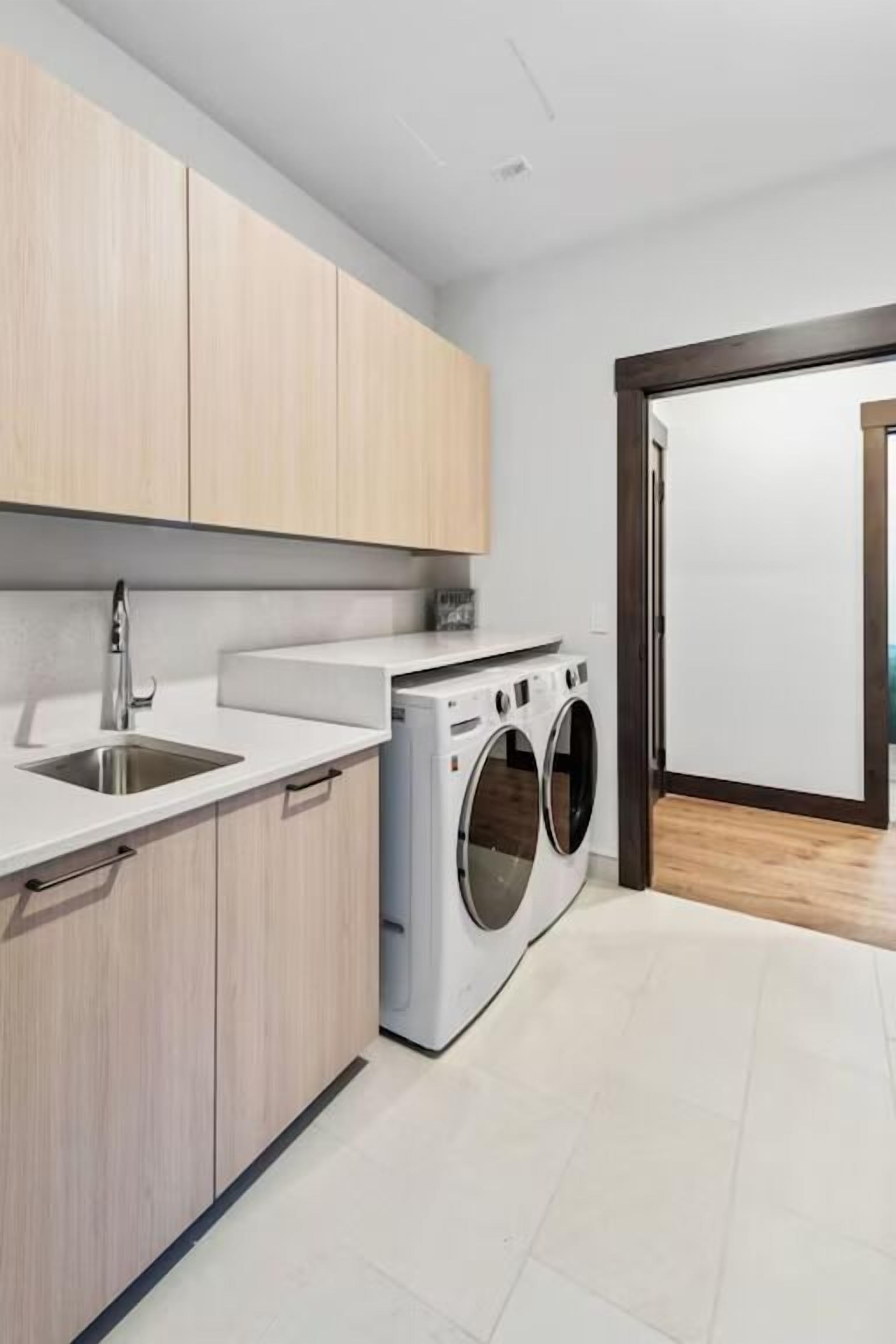 A modern kitchen with light wooden cabinets, a white countertop, a stainless steel sink, and a front-loading washing machine.