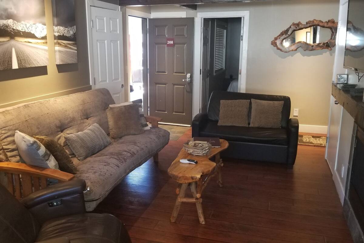 Ein Wohnzimmer mit braunem Ledersofa, einem schwarzen Lederstuhl und einem Couchtisch aus Holz. Eine Tür mit der Nummer 208, ein Spiegel an der Wand und ein Fernseher auf der rechten Seite sind ebenfalls zu sehen.