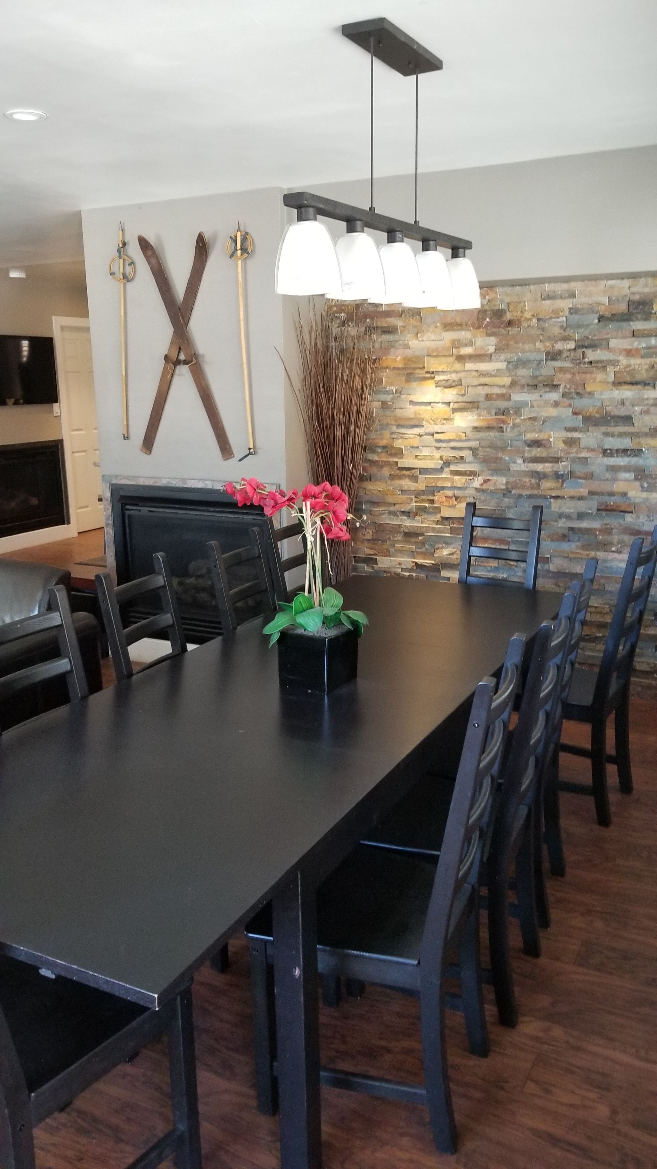 A dining area with a long table, black chairs, a modern chandelier, a stone wall, and a decorative plant with red flowers.
