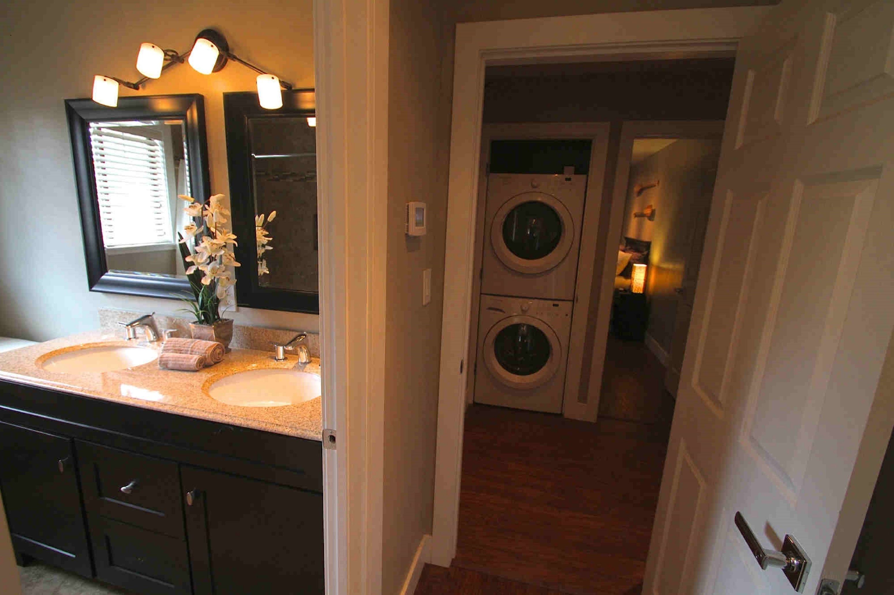 A bathroom with two sinks, a mirror, and a towel rack. There is a door leading to a laundry area with a washing machine and dryer.